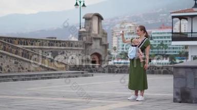 一个带着婴儿的年轻女人去欧洲旅行。 站在海边的广场上打电话